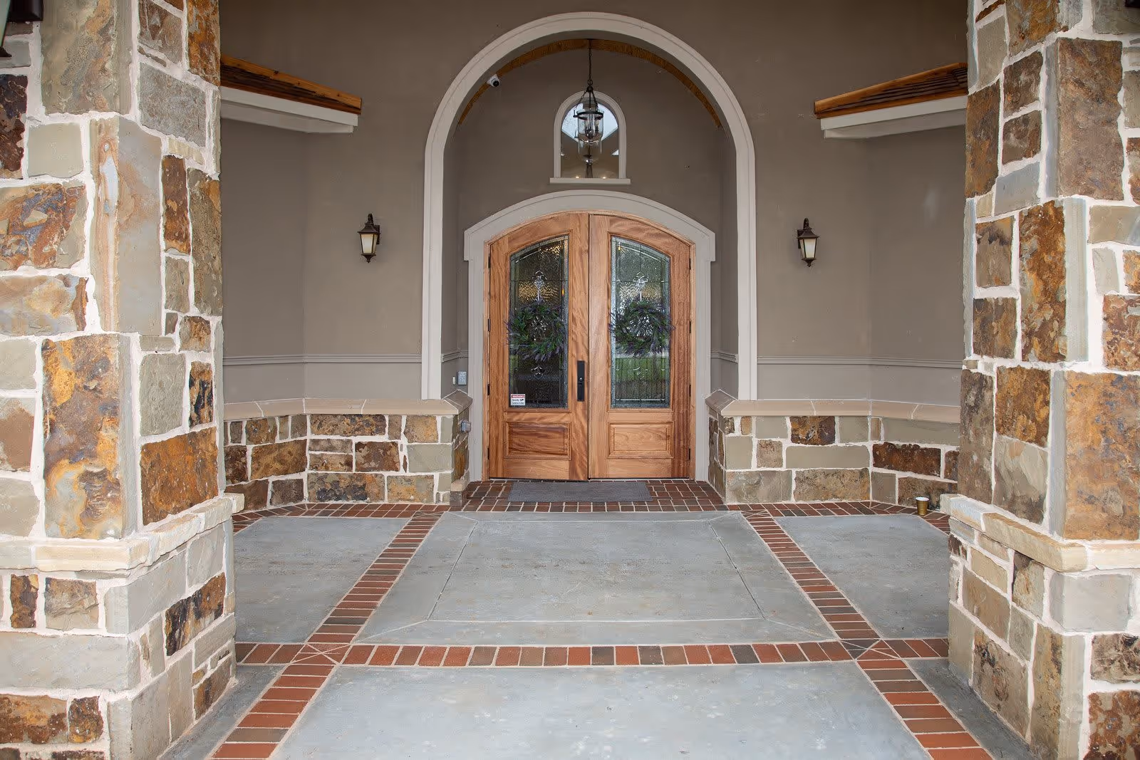 Stone and brick entranceway with two wooden double doors featuring decorative glass panels and wreaths, flanked by stone columns and wall-mounted lantern lights.