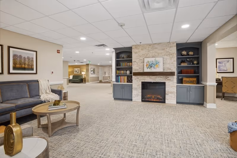 A spacious and well-lit common area in a senior living facility featuring a cozy electric fireplace with stone surround and built-in shelves filled with books and decorative items. To the left, there is a blue sofa with a throw blanket and a round wooden coffee table with books on it. The room has beige walls, carpeted flooring, and framed artwork on the walls. The ceiling has recessed lighting and a smoke detector.