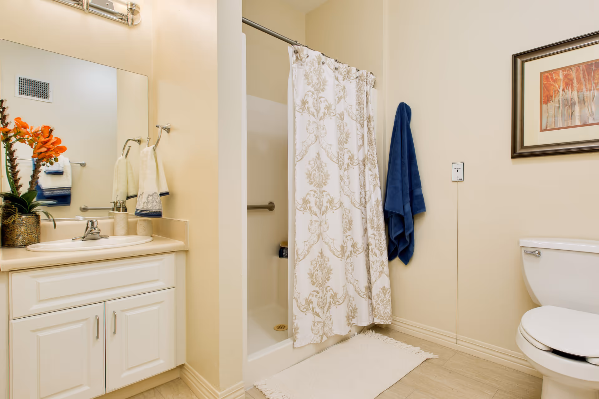 A clean bathroom with a white vanity and sink, a large mirror above the sink, a shower with a decorative curtain, a white toilet, a blue towel hanging on the wall, and a framed artwork. There is also a small plant with orange flowers on the vanity.