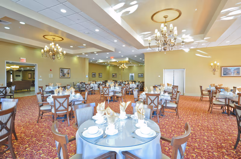 A spacious dining room with multiple round tables covered with light blue tablecloths, each set with white plates, cups, glasses, and beige folded napkins. The room features red patterned carpet, beige walls, several chandeliers hanging from the ceiling, and framed artwork on the walls.