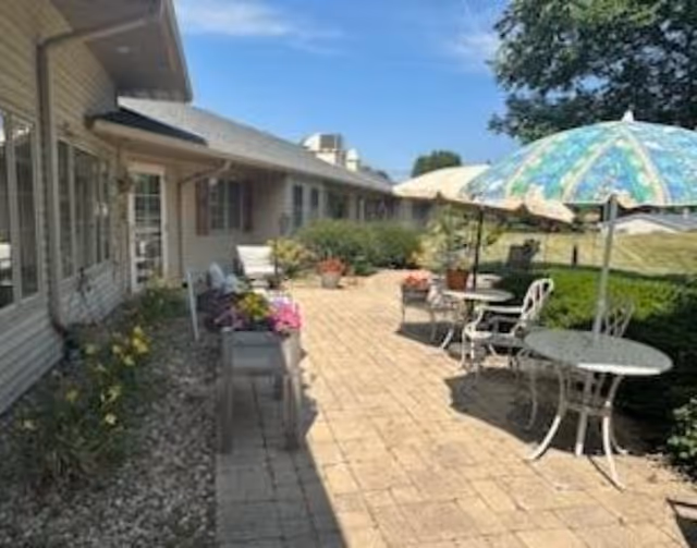 Outdoor patio area at Courtyard Estates of Monmouth with paved walkway, white metal tables and chairs, colorful umbrellas, potted plants, and shrubs alongside a single-story building under a clear blue sky.