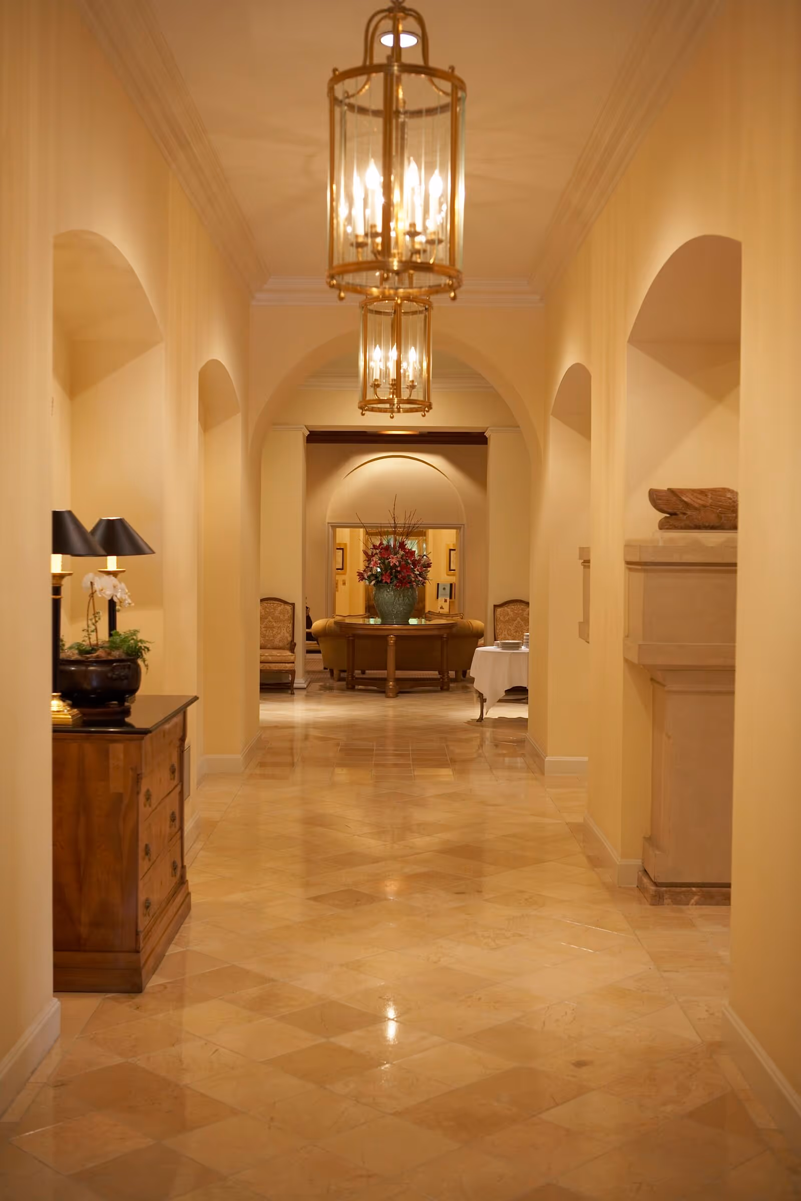Well-lit elegant interior hallway with tiled floor, archways, hanging lanterns, and a seating area with a floral arrangement at the end.