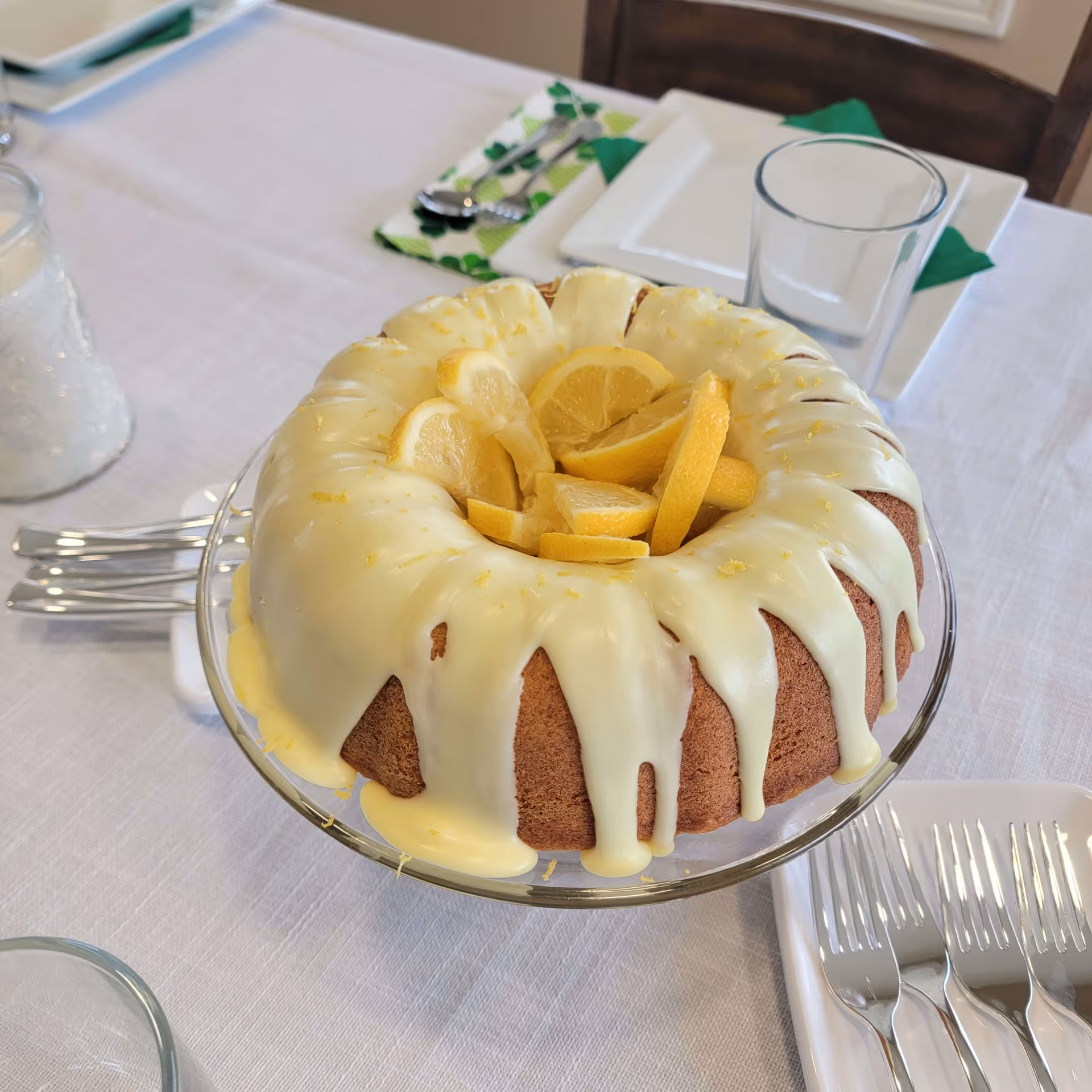 A lemon bundt cake with white icing and lemon slices on top, placed on a glass cake stand on a white tablecloth. Surrounding the cake are plates, forks, glasses, and green and white napkins with silverware.