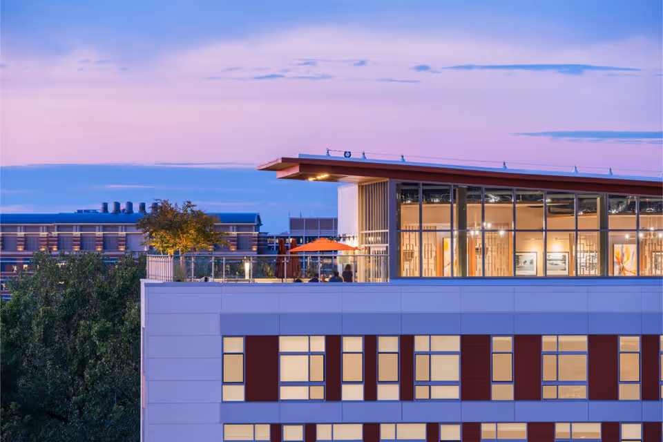 Exterior view of a modern building at dusk with large windows and a rooftop terrace featuring trees and orange umbrellas. The sky is a gradient of purple and blue with some clouds.