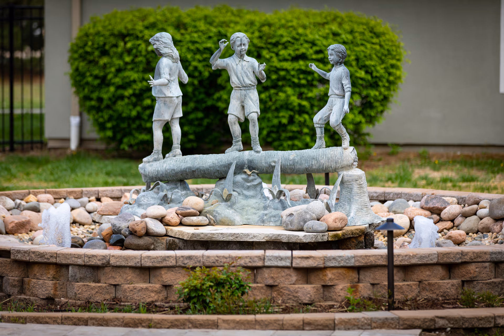 A stone sculpture of three children playing on a log, situated in the center of a circular stone water fountain with small water jets and surrounded by rocks and greenery.
