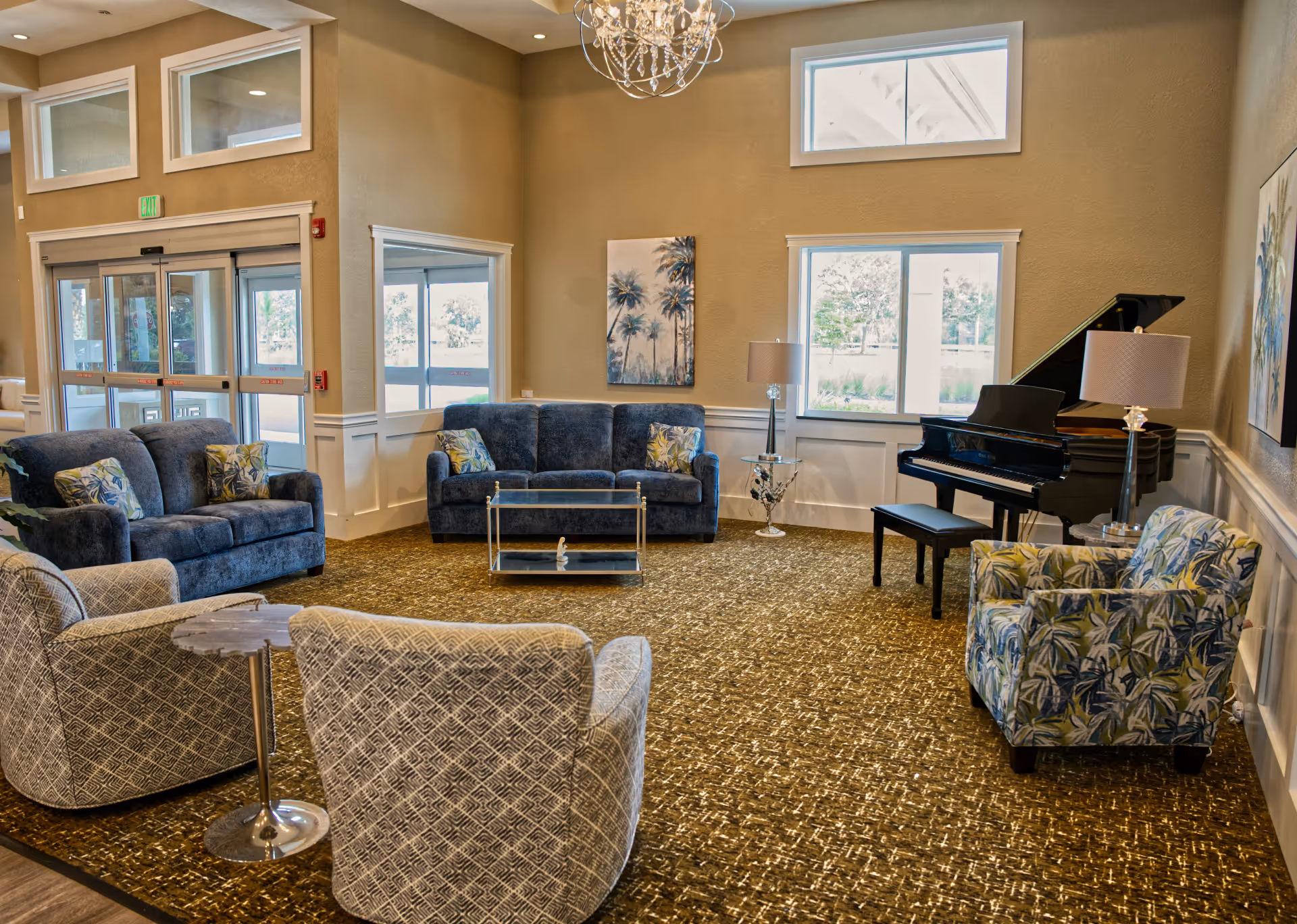 Spacious senior living lounge with blue sofas, patterned chairs, piano, and chandelier.