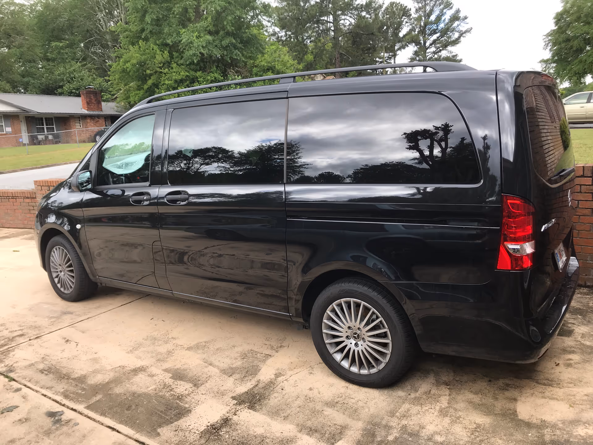 A black Mercedes-Benz van parked on a concrete driveway in front of a brick wall with trees and a house in the background.