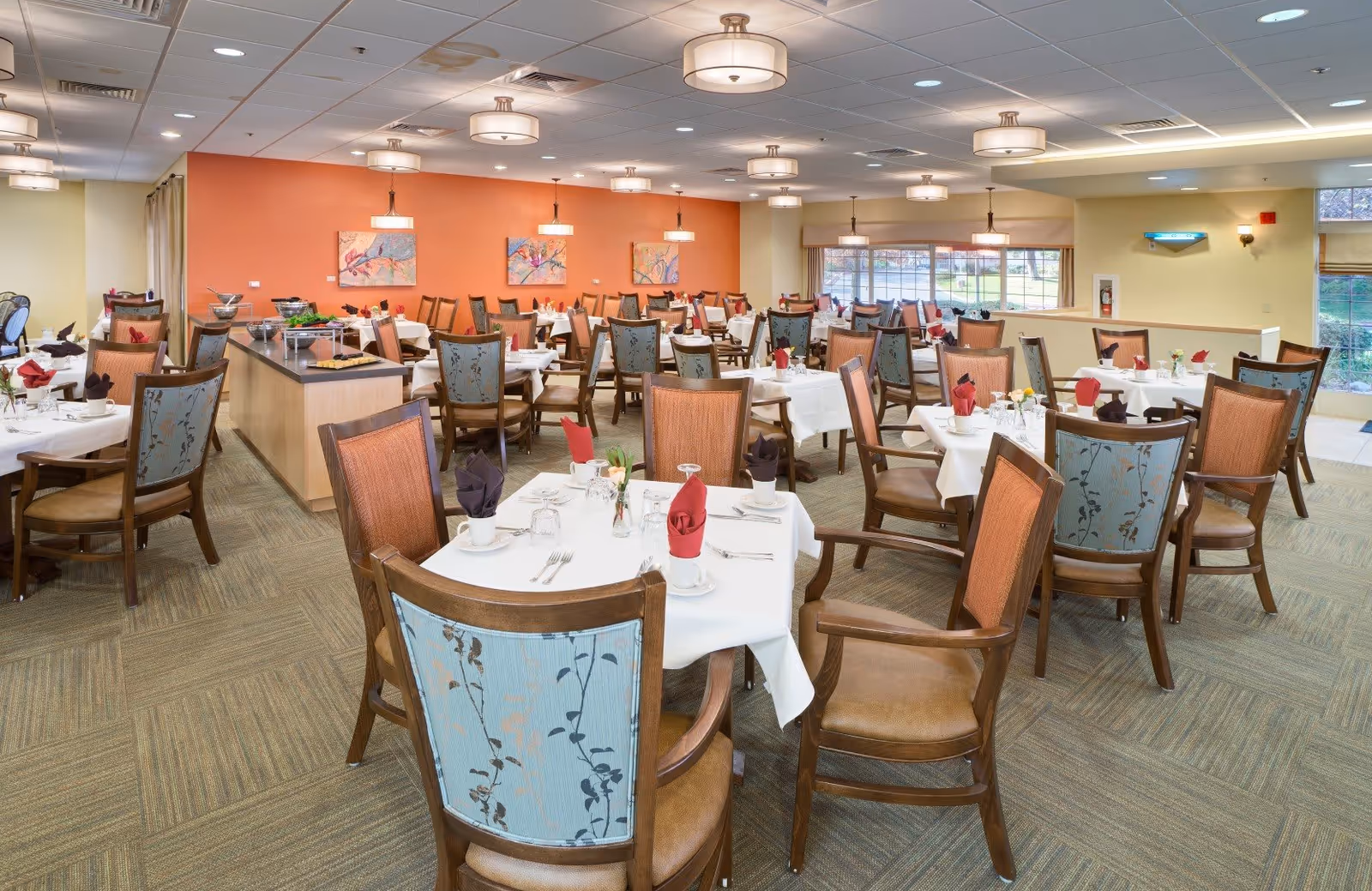A spacious dining room with multiple tables covered in white tablecloths, each set with cups, glasses, silverware, and folded red or dark napkins. The chairs have wooden frames with patterned upholstery in blue and orange tones. The room has a warm ambiance with orange and beige walls, ceiling lights, and large windows letting in natural light.
