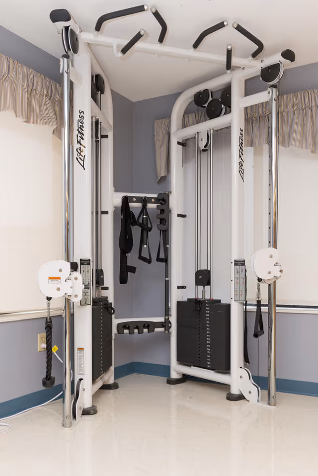A Life Fitness multi-station gym machine in a corner of a room with light-colored walls, windows with striped valances, and a tiled floor.