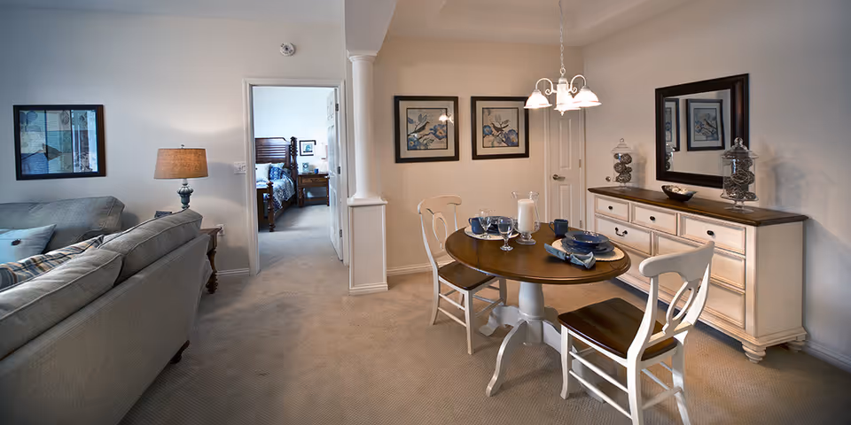 A cozy interior space featuring a small round dining table set for two with blue dishes and glassware, two white wooden chairs, a sideboard with decorative jars and a large mirror above it, a beige carpeted floor, and a glimpse into a bedroom with a wooden bed frame. The living area includes a gray sofa and a lamp on a side table, with framed artwork on the walls.