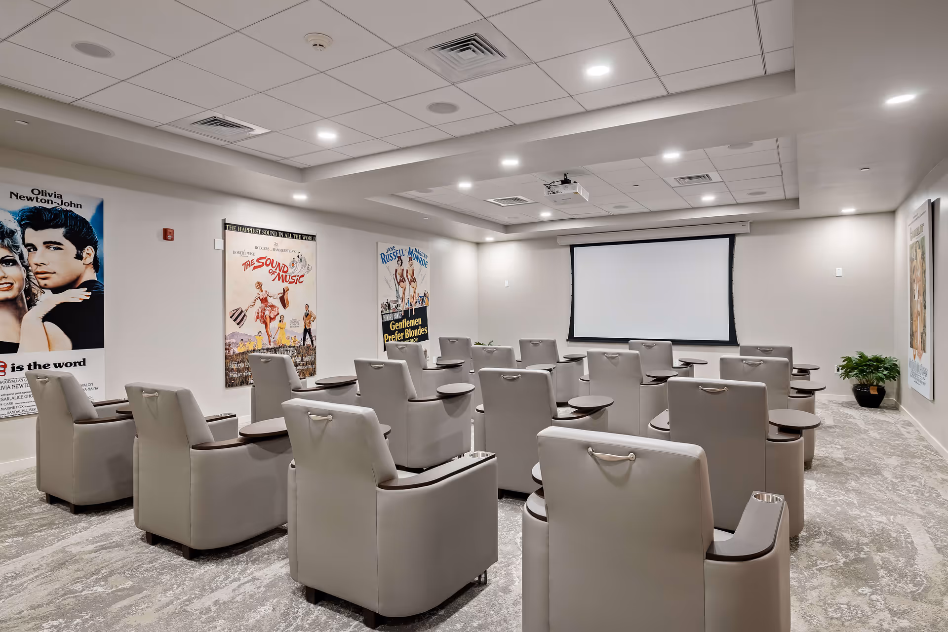 A small theater room with rows of beige cushioned chairs equipped with small attached tables, facing a large white projection screen. The walls are decorated with vintage movie posters including 'Grease', 'The Sound of Music', and 'Gentlemen Prefer Blondes'. The room has a carpeted floor, recessed lighting in the ceiling, and a projector mounted above.