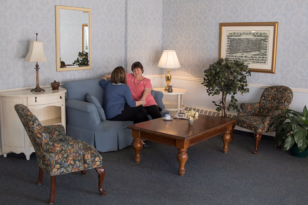 Two women sitting and talking on a blue couch in a cozy living room with floral armchairs, a wooden coffee table, a white side table with a lamp, a framed picture on the wall, and indoor plants.