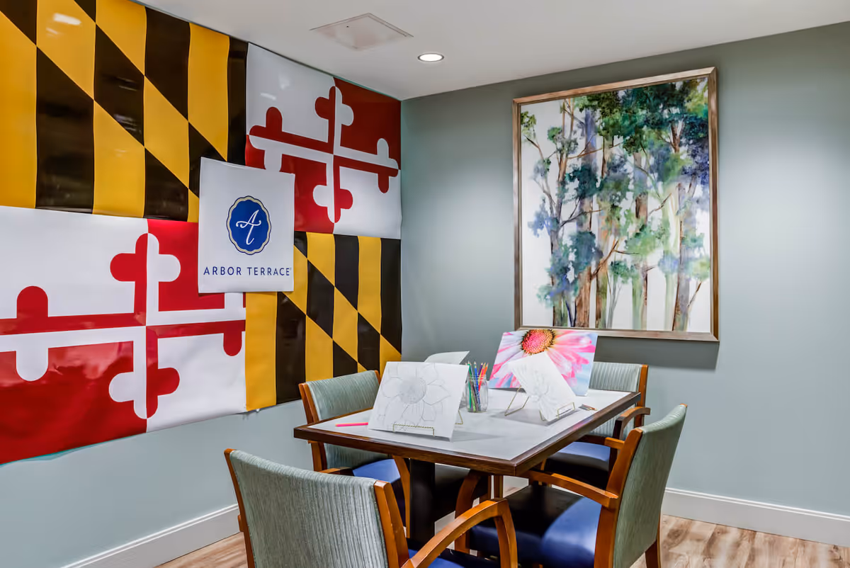 A small activity room with a table and four chairs. On the table are coloring pages with flower outlines and colored pencils in a glass jar. The wall features a large Maryland state flag and a sign with the Arbor Terrace logo. Another wall has a framed painting of trees.