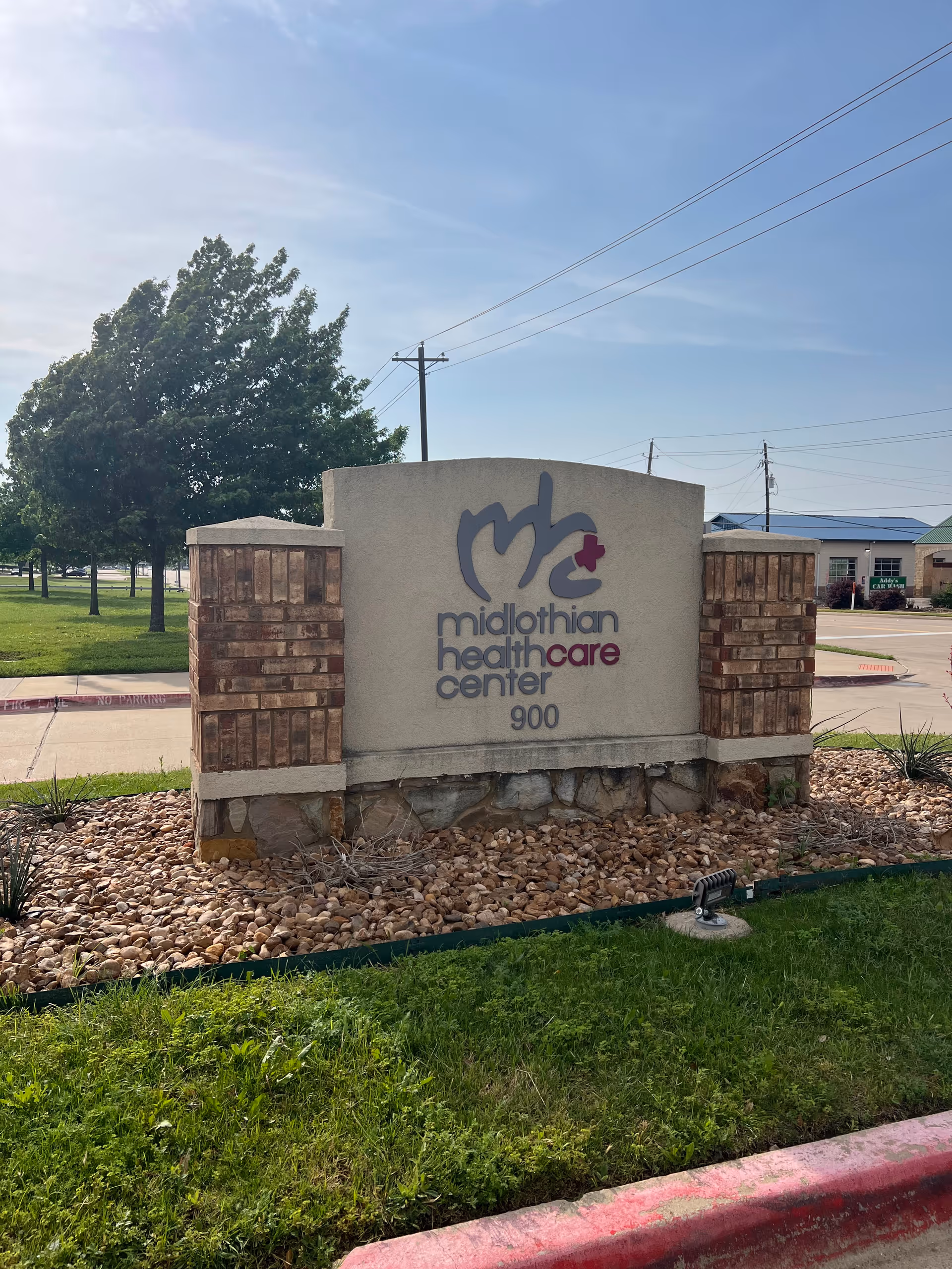 Outdoor view of a sign for Midlothian Healthcare Center, featuring the center's logo and address number 900. The sign is made of brick and concrete, surrounded by rocks and grass, with trees and a clear sky in the background.