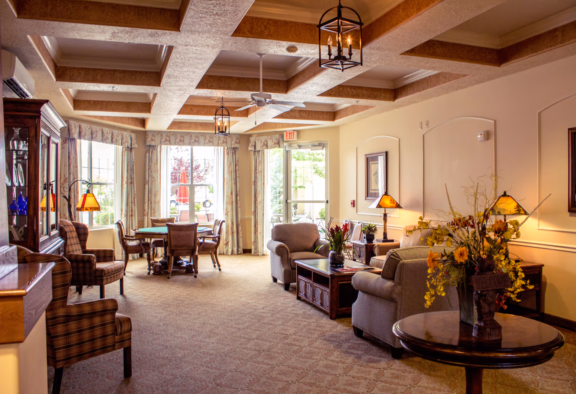 A cozy and well-lit living room area in a senior living facility with comfortable armchairs, a coffee table, a round table with chairs near large windows, decorative lamps, floral arrangements, and a ceiling with wooden beams.