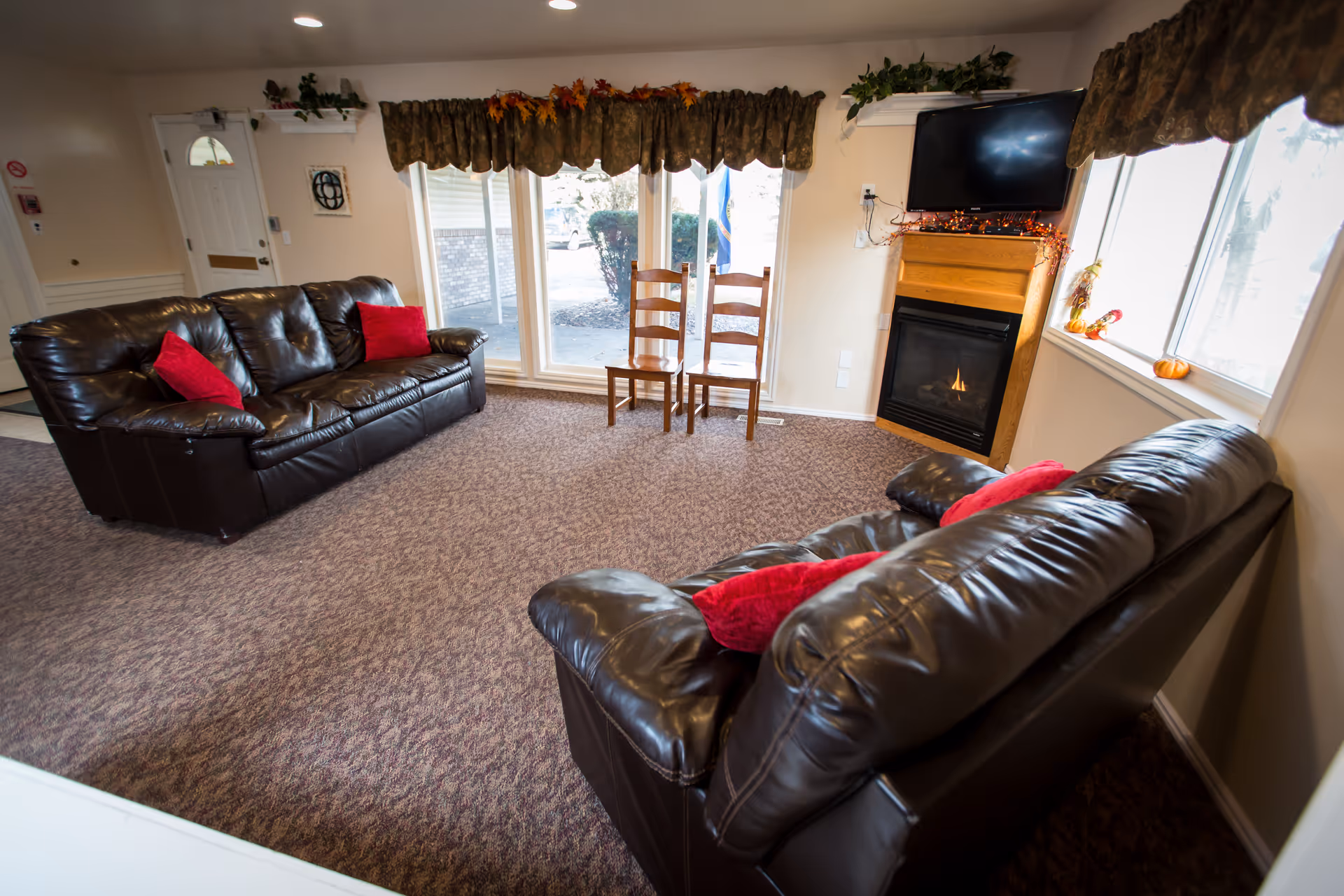 A cozy living room with two dark brown leather sofas adorned with red pillows, a fireplace with a TV mounted above it, two wooden chairs near large windows with floral valances, and a carpeted floor.