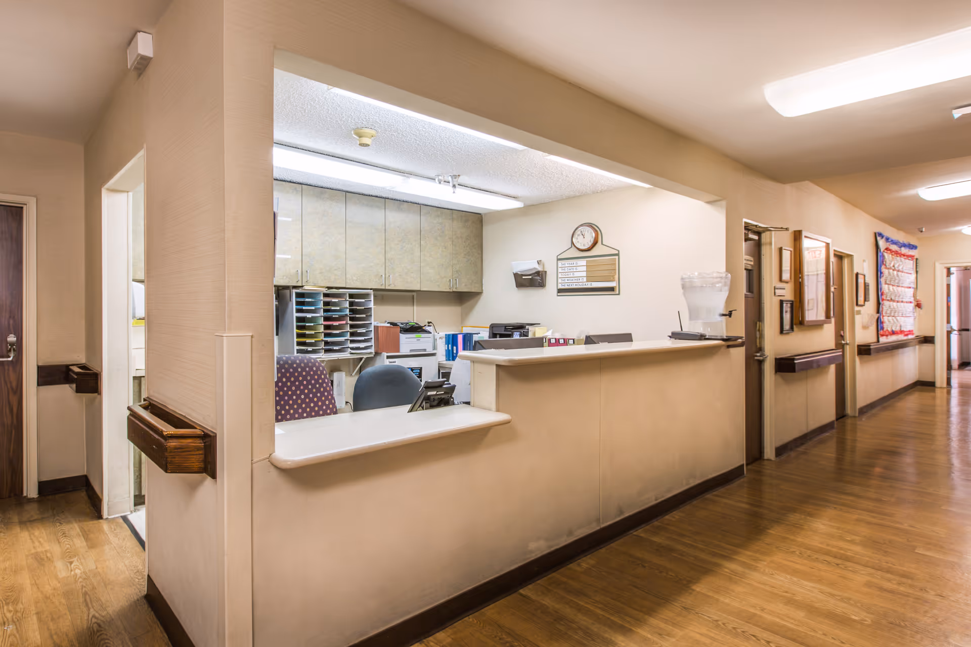 Reception desk area inside The Earlwood facility with office chairs, filing shelves, a clock on the wall, and a water dispenser. The hallway adjacent to the reception desk has wooden floors, handrails, and doors leading to other rooms.