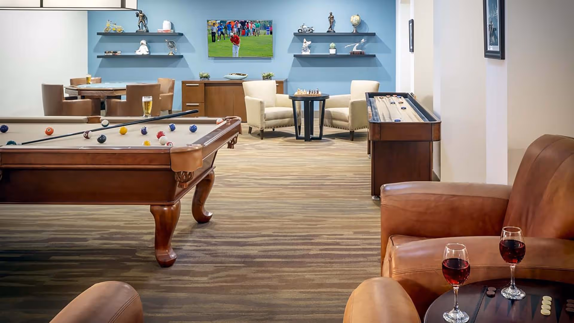 A recreational room featuring a pool table with billiard balls and cues, a shuffleboard table, two beige armchairs around a small table with a chess set, a wall-mounted TV showing a golf game, and a seating area with two leather armchairs and a small table holding two glasses of red wine.