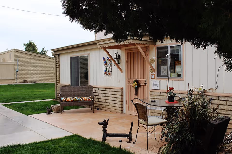 Outdoor patio area of a building with a small tiled seating area featuring a wicker bench with a colorful cushion, a round glass table with two chairs, and various plants. The building has a beige and brick exterior with a peach-colored door and window. A tree partially shades the patio.