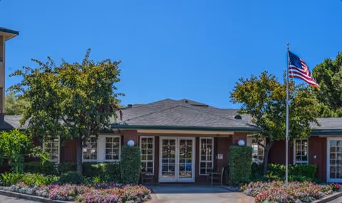 Front exterior view of Carmel Village Retirement Community building with a gray roof, brick walls, multiple windows, and a central entrance with glass doors. The entrance is flanked by green shrubs and flowering plants, with an American flag on a flagpole to the right. The sky is clear and blue.