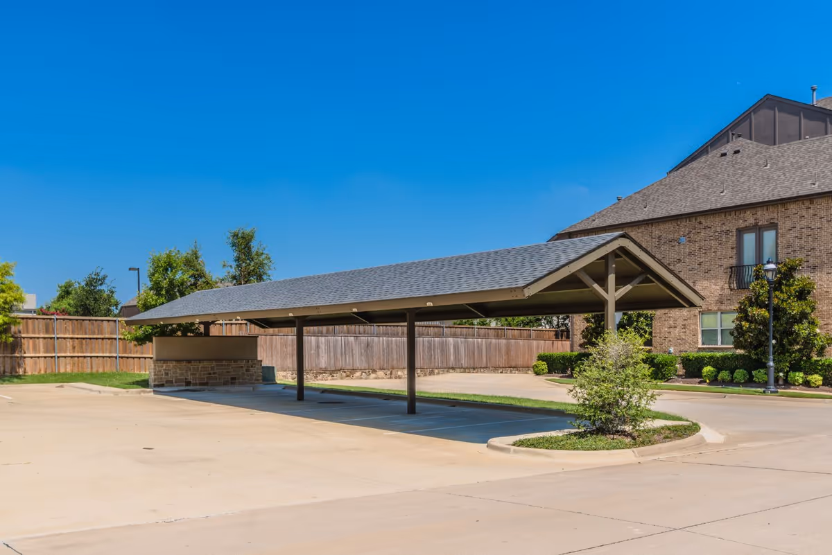 Outdoor covered parking area with a pitched roof supported by metal posts, adjacent to a brick building with windows and landscaping including bushes and a small tree, under a clear blue sky.