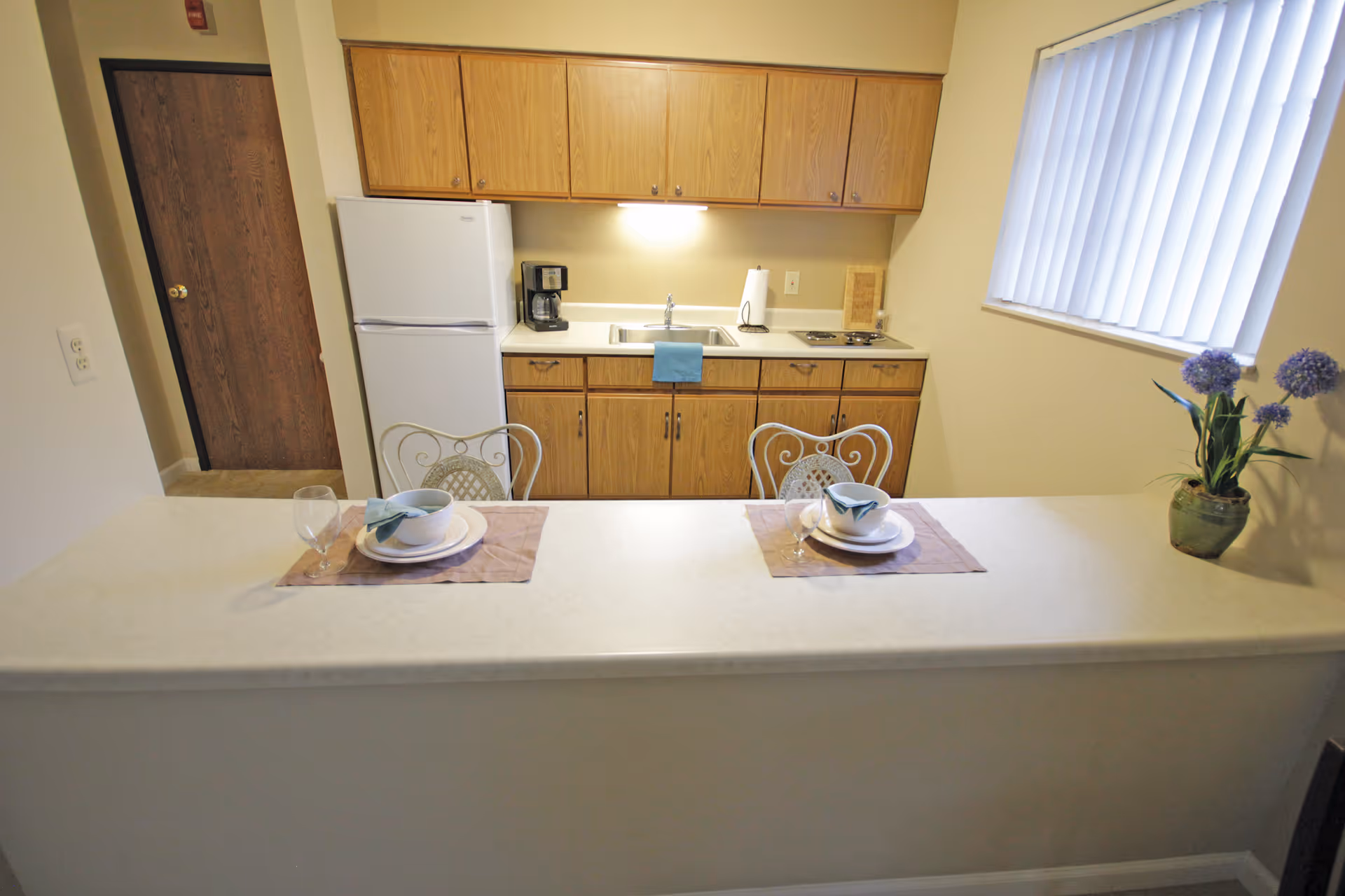 A small kitchen area with wooden cabinets, a white refrigerator, a coffee maker, a sink, and a two-burner stove. In front of the kitchen is a counter with two place settings, each with a bowl, plate, napkin, and glass. There are two decorative metal chairs at the counter and a window with vertical blinds on the right side. A potted plant with purple flowers is on the counter near the window.