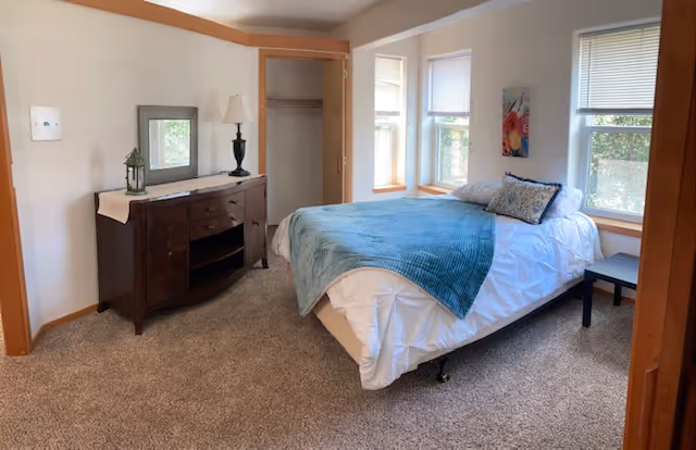 A cozy bedroom with a bed covered in white bedding and a teal throw blanket, a wooden dresser with a lamp and decorative items, a mirror on the wall, an open closet, and three windows letting in natural light.