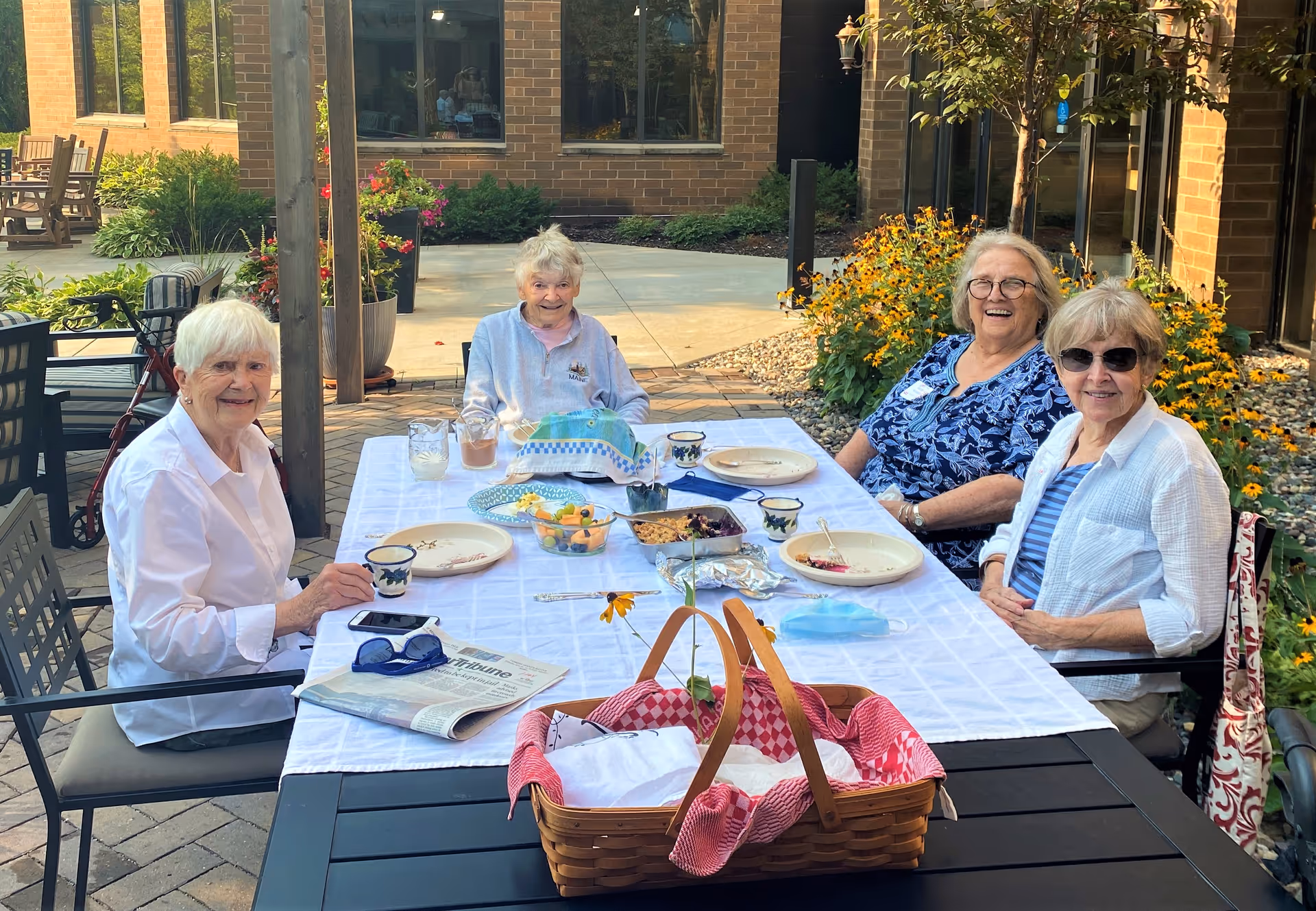 Four elderly women sitting around a rectangular outdoor table covered with a white tablecloth, enjoying a meal together. The table has plates, cups, a basket with a red and white cloth, and some food dishes. The setting is a patio area with brick walls, windows, plants, and flowers in the background. The women are smiling and appear to be having a pleasant time.