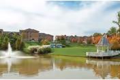 Pond with a fountain, a white gazebo, and landscaped lawns in front of senior living buildings under a partly cloudy sky.