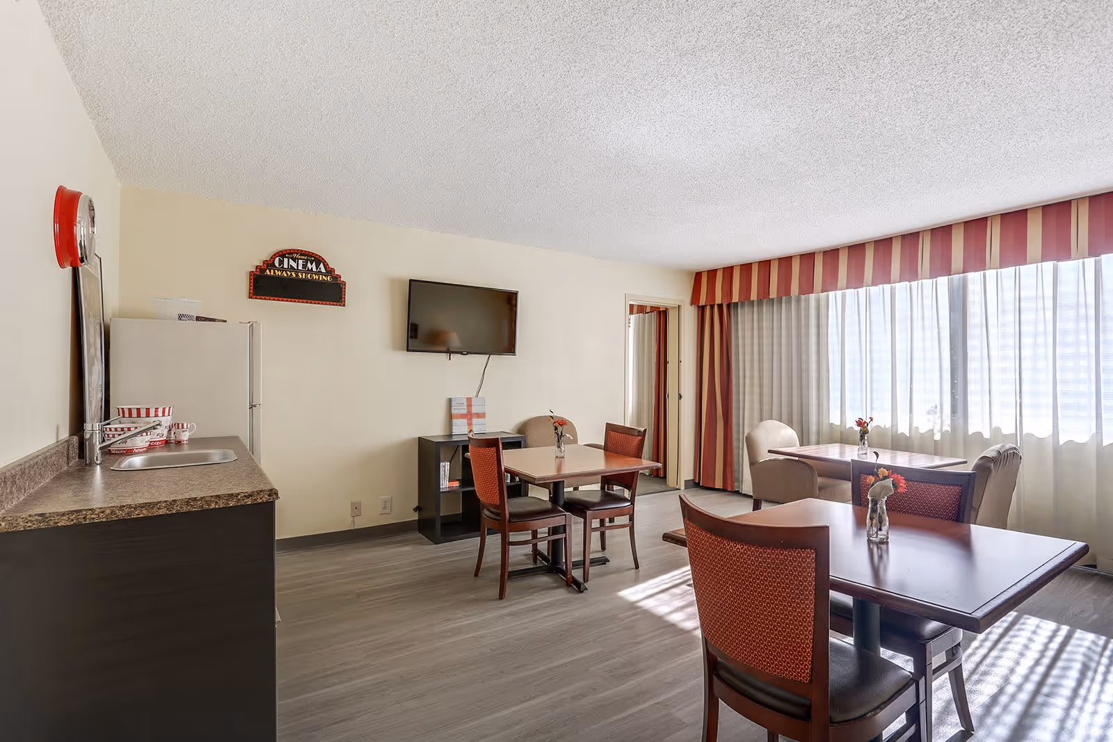 A bright common area in a senior living facility with several tables and chairs arranged for dining or socializing. There is a small kitchenette with a sink and countertop on the left side, a wall-mounted flat-screen TV, and a decorative sign that reads 'Cinema Always Showing'. Large windows with sheer curtains and striped valances allow natural light to fill the room.