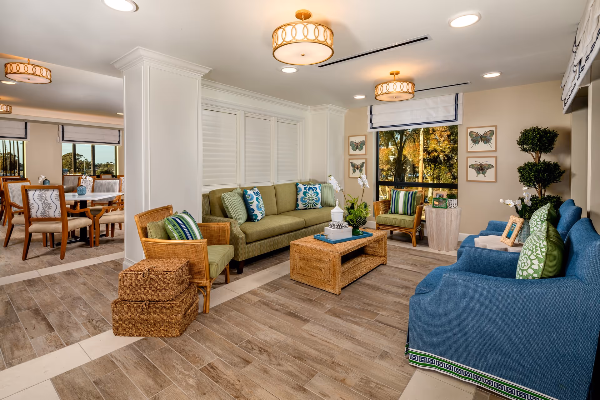 Bright furnished common living room area with green and blue sofas, wicker chairs, a coffee table, and a dining area visible in the background.