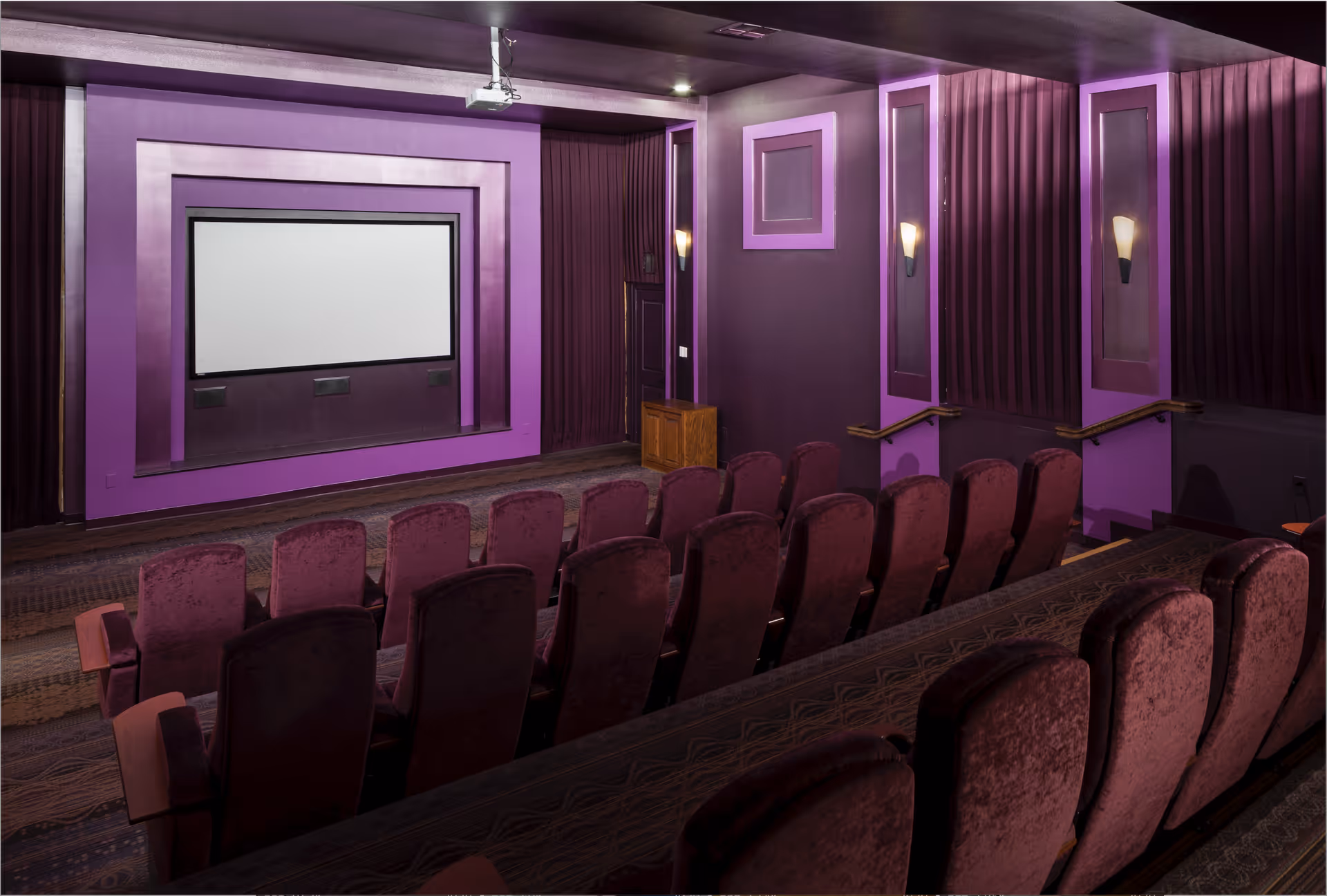 Rows of plush purple theater seats facing a blank screen in a small movie theater with purple-accented walls.