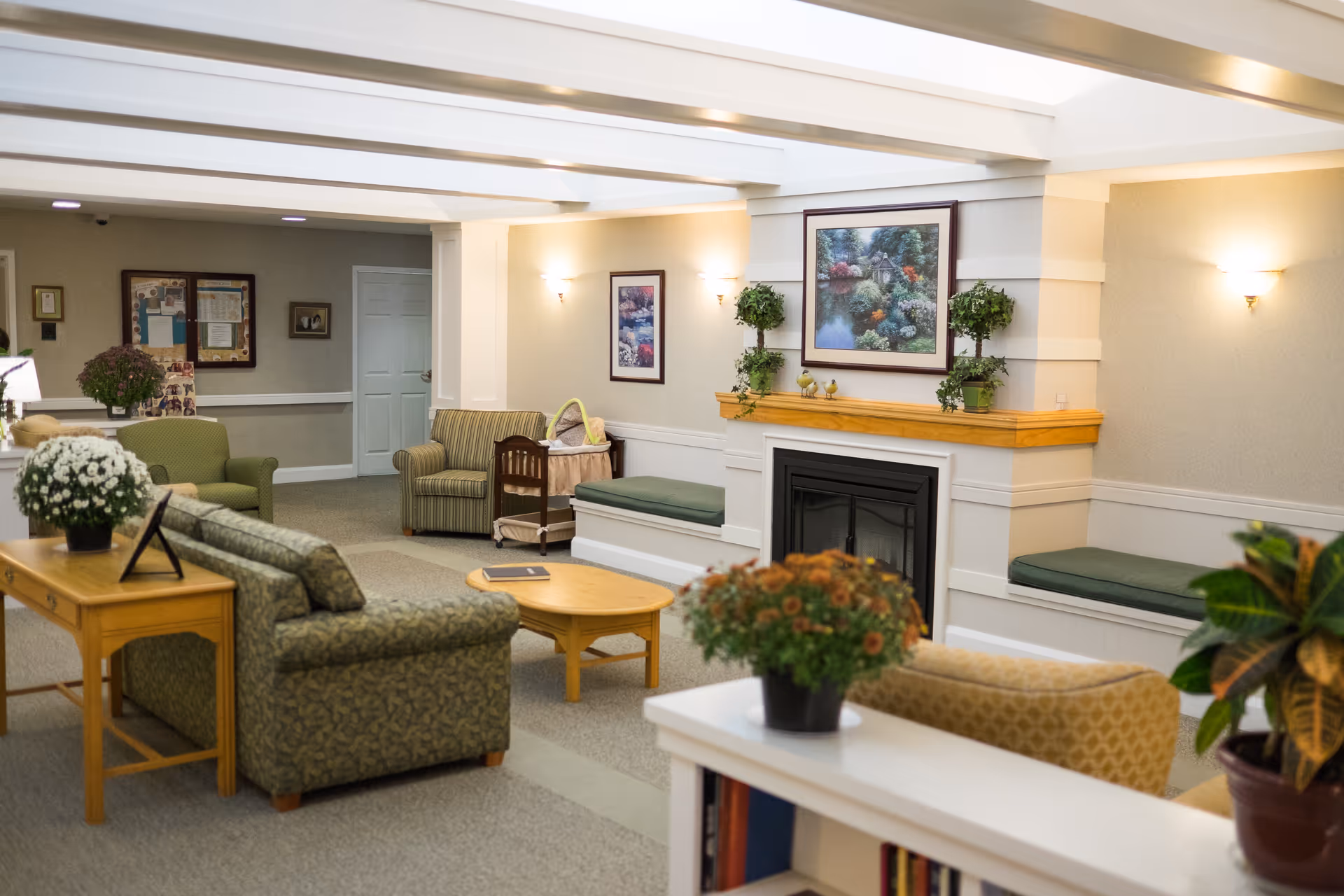 A cozy living room area in a senior living facility with patterned sofas, armchairs, a wooden coffee table, and side tables adorned with potted plants and flowers. The room features a fireplace with a wooden mantle decorated with small plants and a framed painting above it. The walls have soft lighting fixtures, and there are cushioned bench seats along the wall.