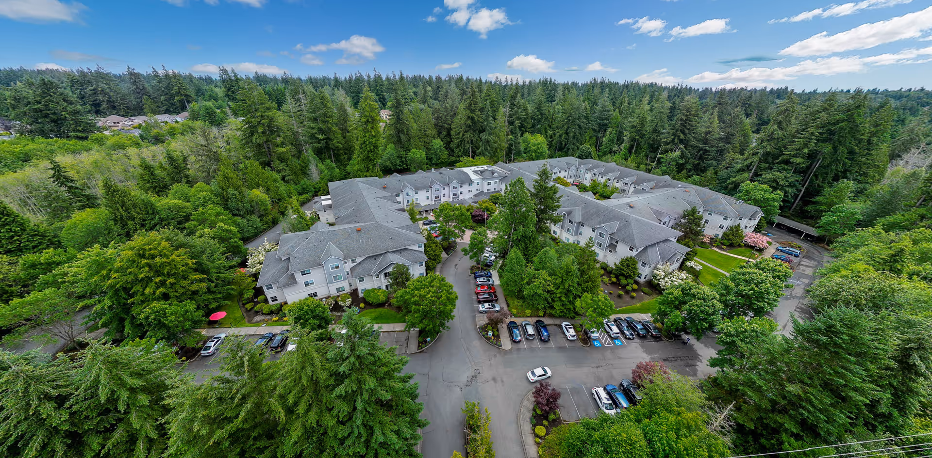 Aerial view of Cogir of Mill Creek senior living facility surrounded by dense green trees and forest. The facility consists of multiple connected buildings with gray roofs, parking lots with several cars, and well-maintained landscaping. The sky is blue with scattered clouds.