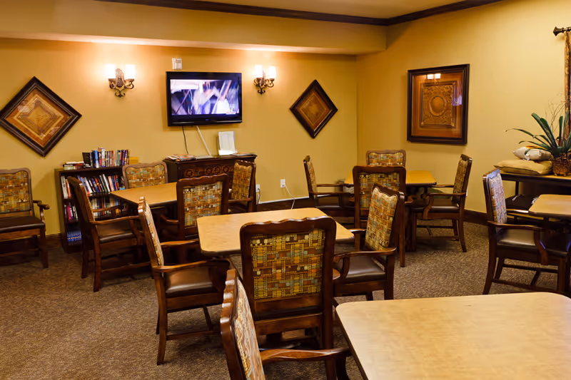 A cozy common room with several wooden tables and cushioned chairs arranged around the room. The walls are painted a warm beige color and decorated with framed artwork. A flat-screen TV is mounted on the wall above a small cabinet, with two wall sconces providing soft lighting. A bookshelf filled with books is visible in the corner, and a table with decorative plants and cushions is on the right side.