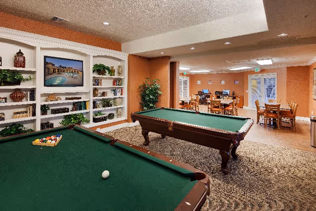 Interior view of a senior living facility's common area featuring two pool tables in the foreground, a built-in white bookshelf with decorative items and a TV mounted above it on the left wall, and a seating area with tables and chairs in the background. The walls are painted in warm tones and the floor is carpeted near the pool tables and tiled near the seating area.