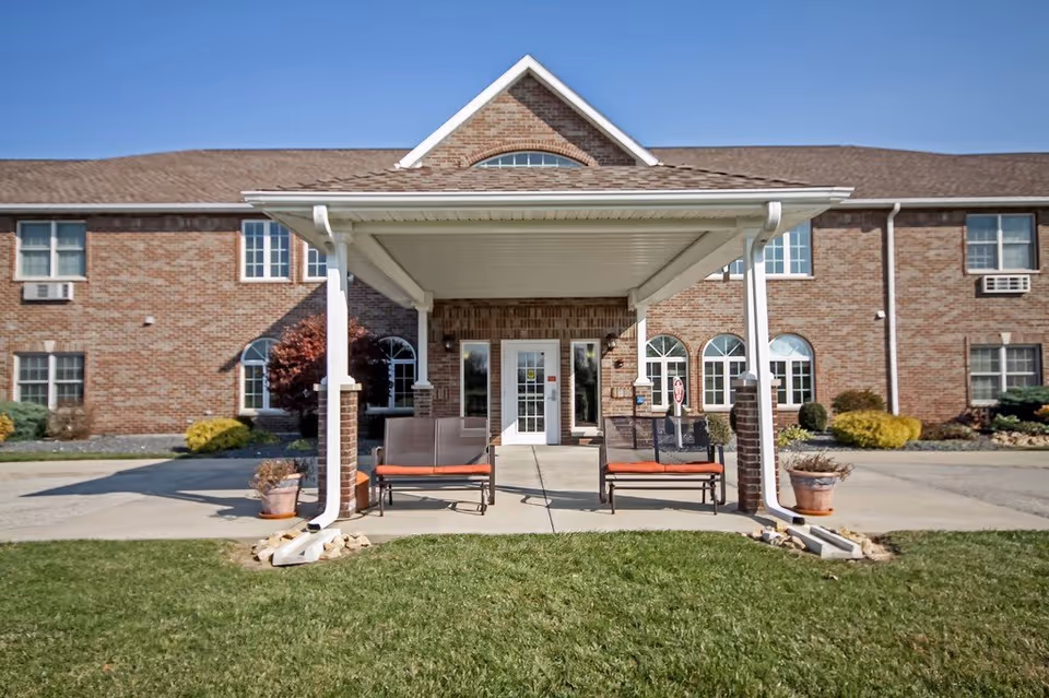 Front entrance of a brick building with a covered porch area featuring two benches with orange cushions. There are potted plants on either side of the entrance, and the building has multiple windows with white frames under a clear blue sky.