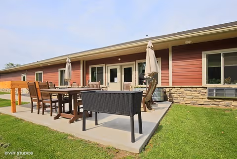 Outdoor patio area with multiple wooden tables and chairs, some with closed umbrellas, adjacent to a single-story building with red siding and stone accents under a partly cloudy sky.