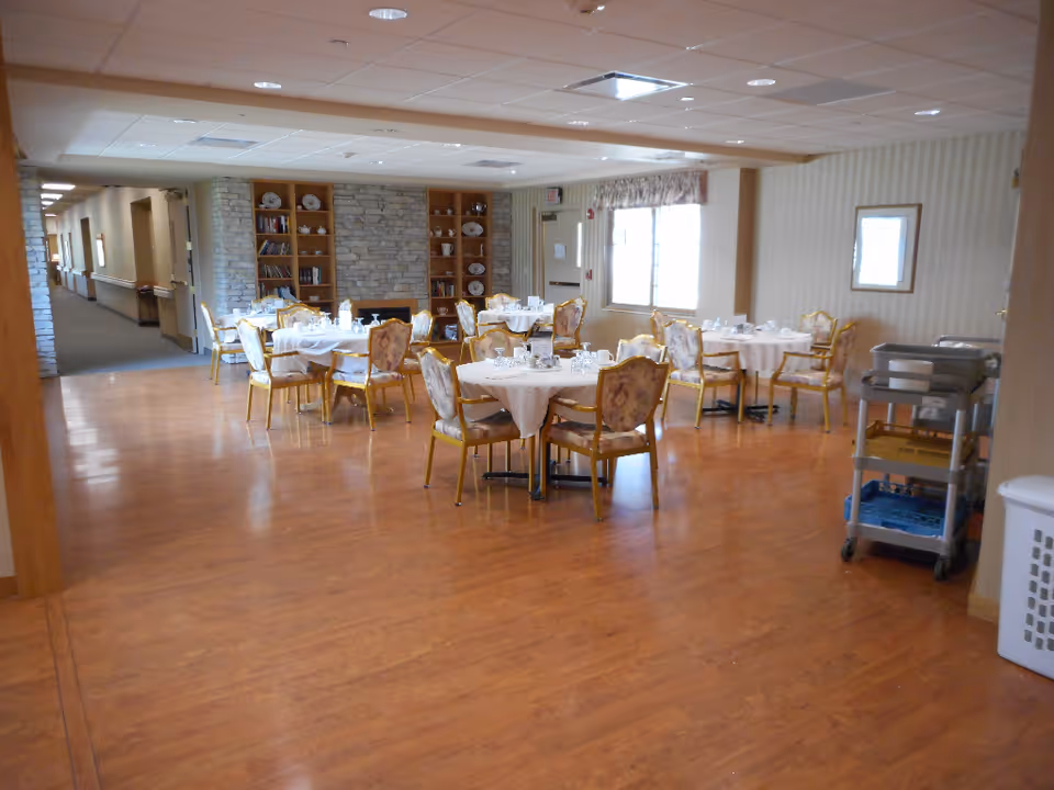 A dining area in a senior living facility with round tables covered with white tablecloths and set with glassware and plates. The room has wooden flooring, upholstered chairs with floral patterns, a window with curtains, and built-in shelves with decorative items and books. A hallway is visible to the left, and a serving cart is on the right side of the image.