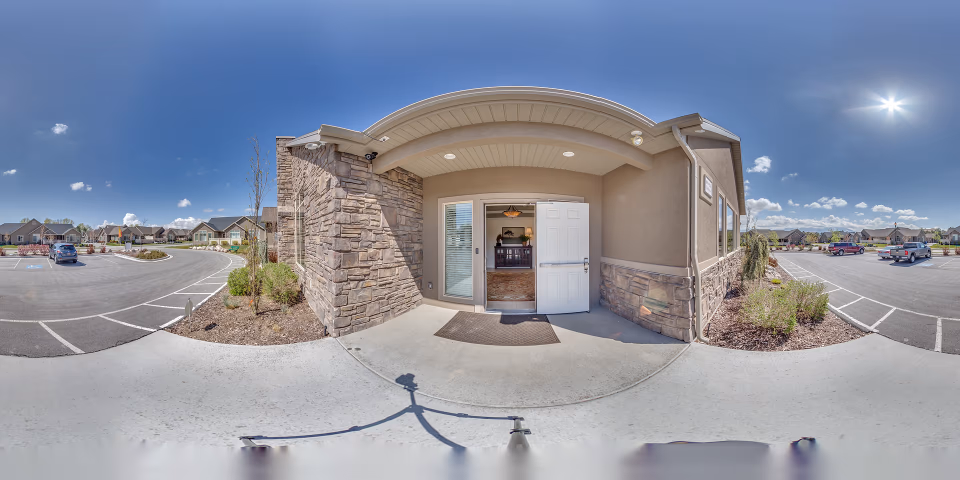 Exterior entrance of Bridlewood Villas facility showing a covered porch with stone and stucco walls, an open white door leading inside, and a parking lot with several cars under a clear blue sky.