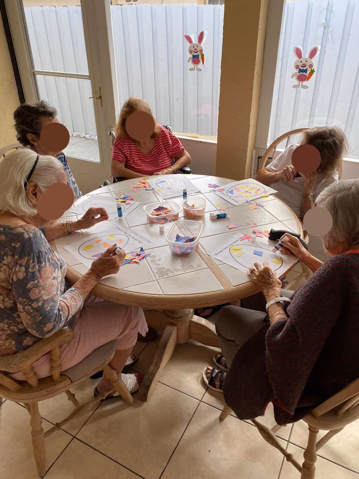 Five elderly women sitting around a round table engaged in a craft activity with colored paper pieces and glue sticks. The room has large windows with bunny decorations on the glass, and the women appear focused on their art projects.