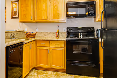 A kitchen with wooden cabinets, a black dishwasher, black electric stove with oven, black microwave above the stove, and a black refrigerator. The countertop is beige with a basket and a bottle on it. The floor has a tiled pattern.