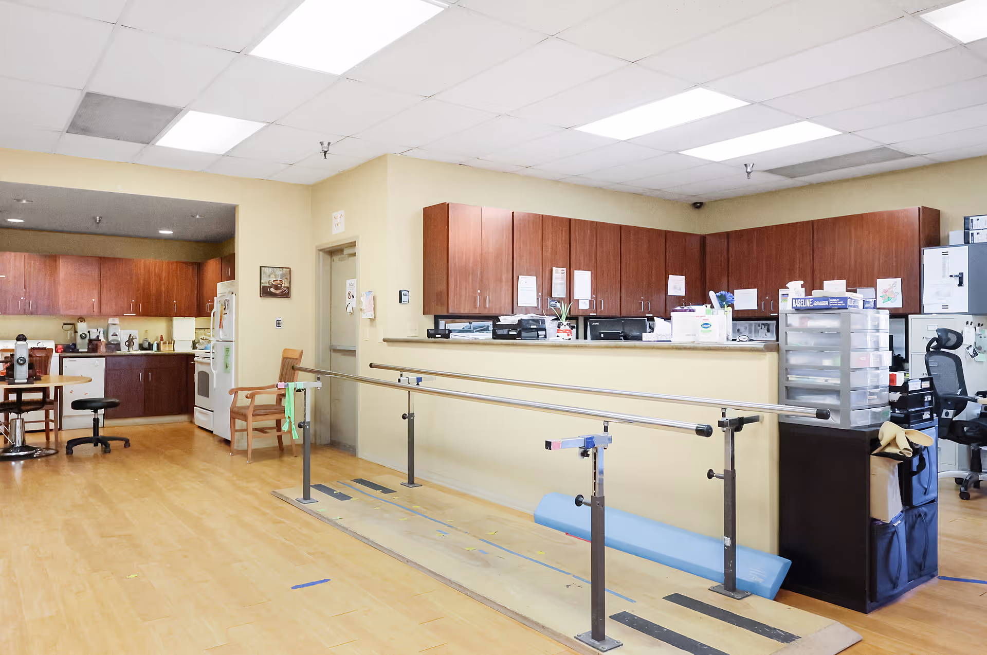 Interior view of a senior living facility area with a kitchen on the left featuring wooden cabinets, a refrigerator, and a stove. In the foreground, there are parallel bars used for physical therapy or rehabilitation. On the right side, there is a reception or work desk with office supplies and chairs behind it. The floor is wooden, and the ceiling has fluorescent lighting.