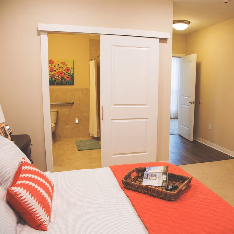 View of a bedroom with a bed covered in white and red bedding, a red and white patterned pillow, and a wicker tray with a book and remote on the bed. A sliding door is partially open, revealing a bathroom with a toilet, grab bar, shower curtain, and a colorful floral painting on the wall. An open door leads to another room with wood flooring and a ceiling light.