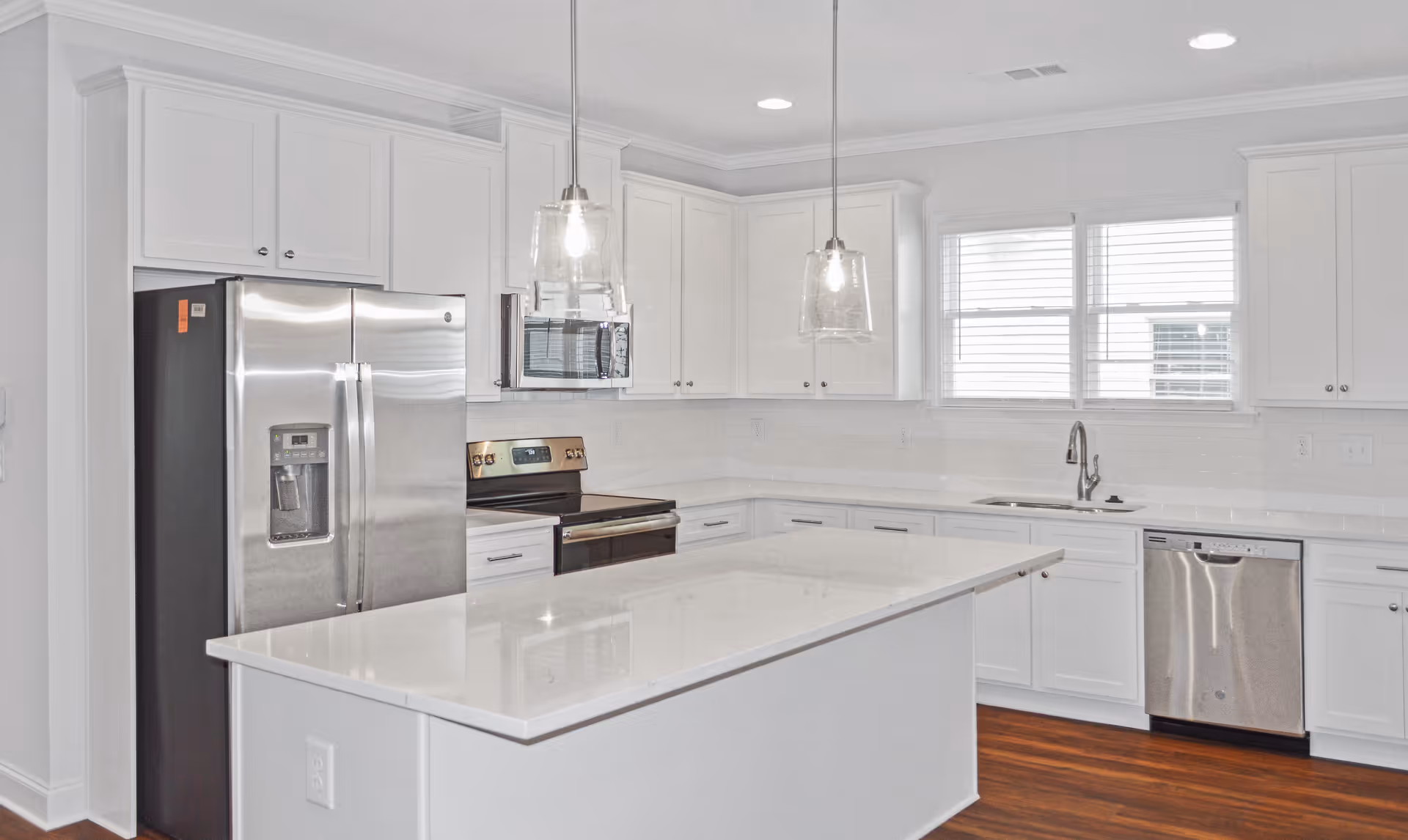 Modern kitchen with white cabinetry, stainless steel refrigerator, oven, microwave, dishwasher, and a large white island countertop. Two pendant lights hang above the island, and there are windows with blinds above the sink.