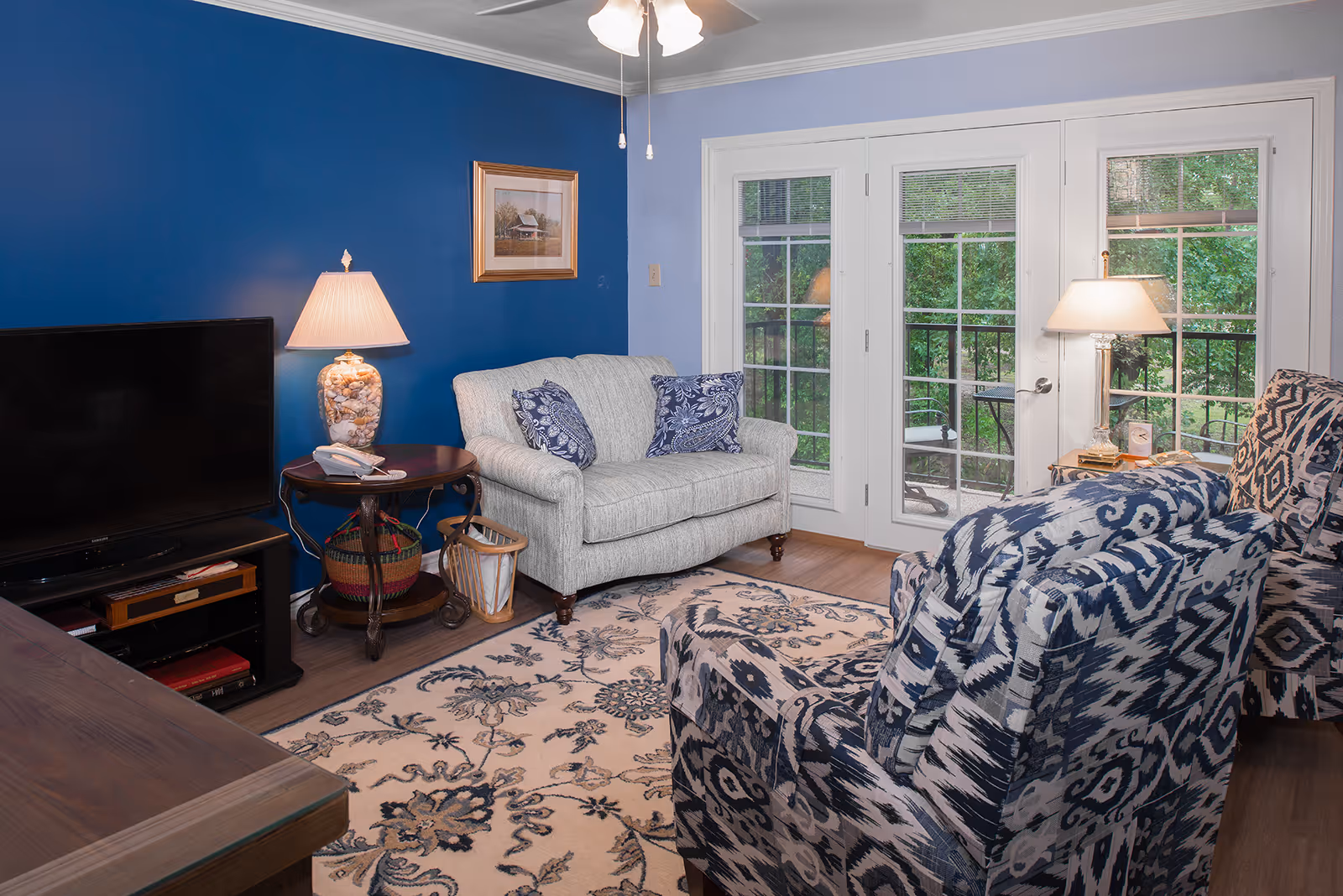 A cozy living room with a blue accent wall, a small loveseat with patterned cushions, two patterned armchairs, a floral area rug, a wooden TV stand with a flat screen TV, two table lamps, and French doors leading to an outdoor area with greenery.