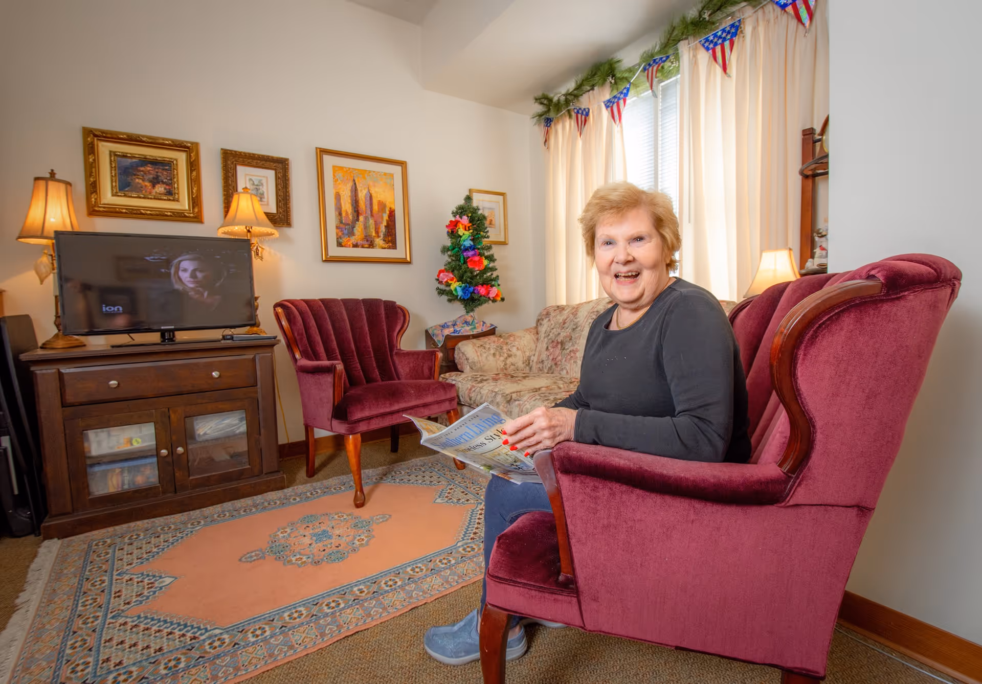An elderly woman sitting in a maroon armchair in a cozy living room, smiling and holding a newspaper. The room features a floral couch, another maroon armchair, a wooden TV stand with a television, framed artwork on the walls, table lamps, and a small decorated Christmas tree near the window with cream-colored curtains and patriotic bunting above.