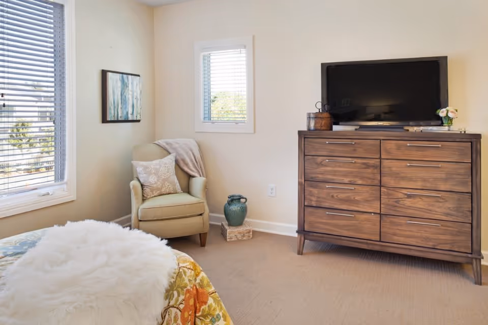 Cozy bedroom corner with an armchair by windows, a wooden dresser topped by a TV, and a partially visible bed.