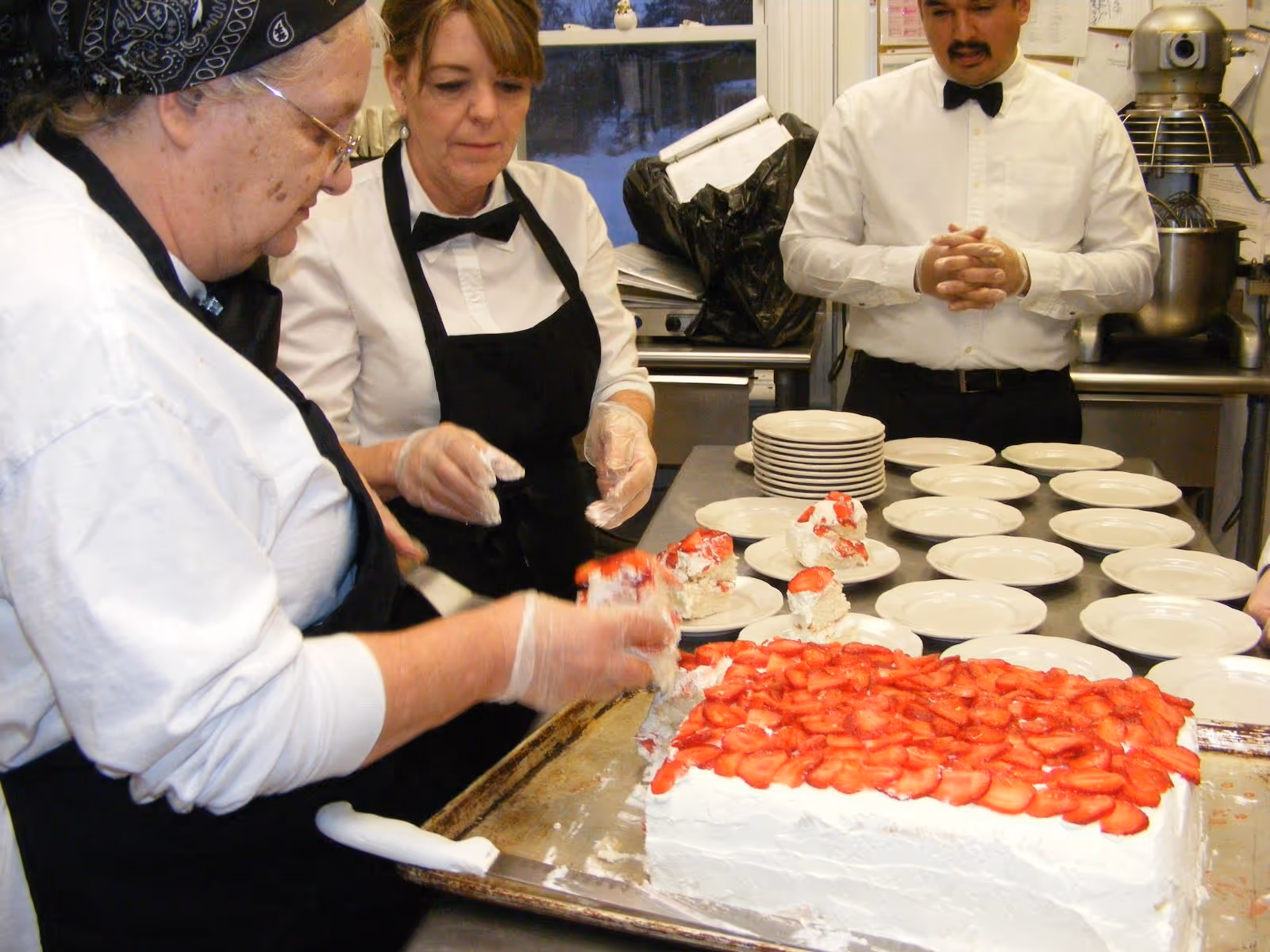 Three kitchen staff members in white shirts, black aprons, and bow ties are preparing and serving slices of a large rectangular cake topped with sliced strawberries onto white plates in a commercial kitchen.