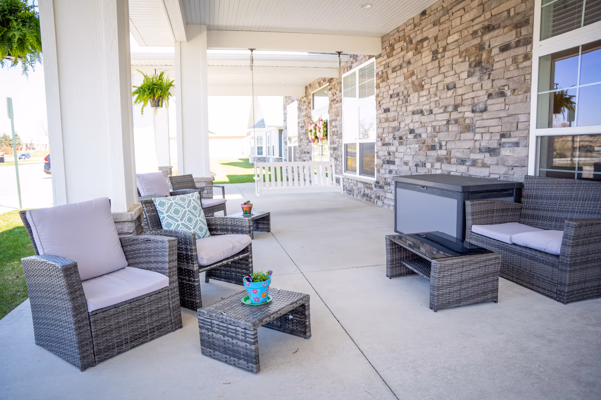 Outdoor covered patio area with wicker furniture including cushioned chairs, a loveseat, and small tables. There are hanging green plants and a white porch swing in the background. The wall is made of stone bricks with windows reflecting the outside view.